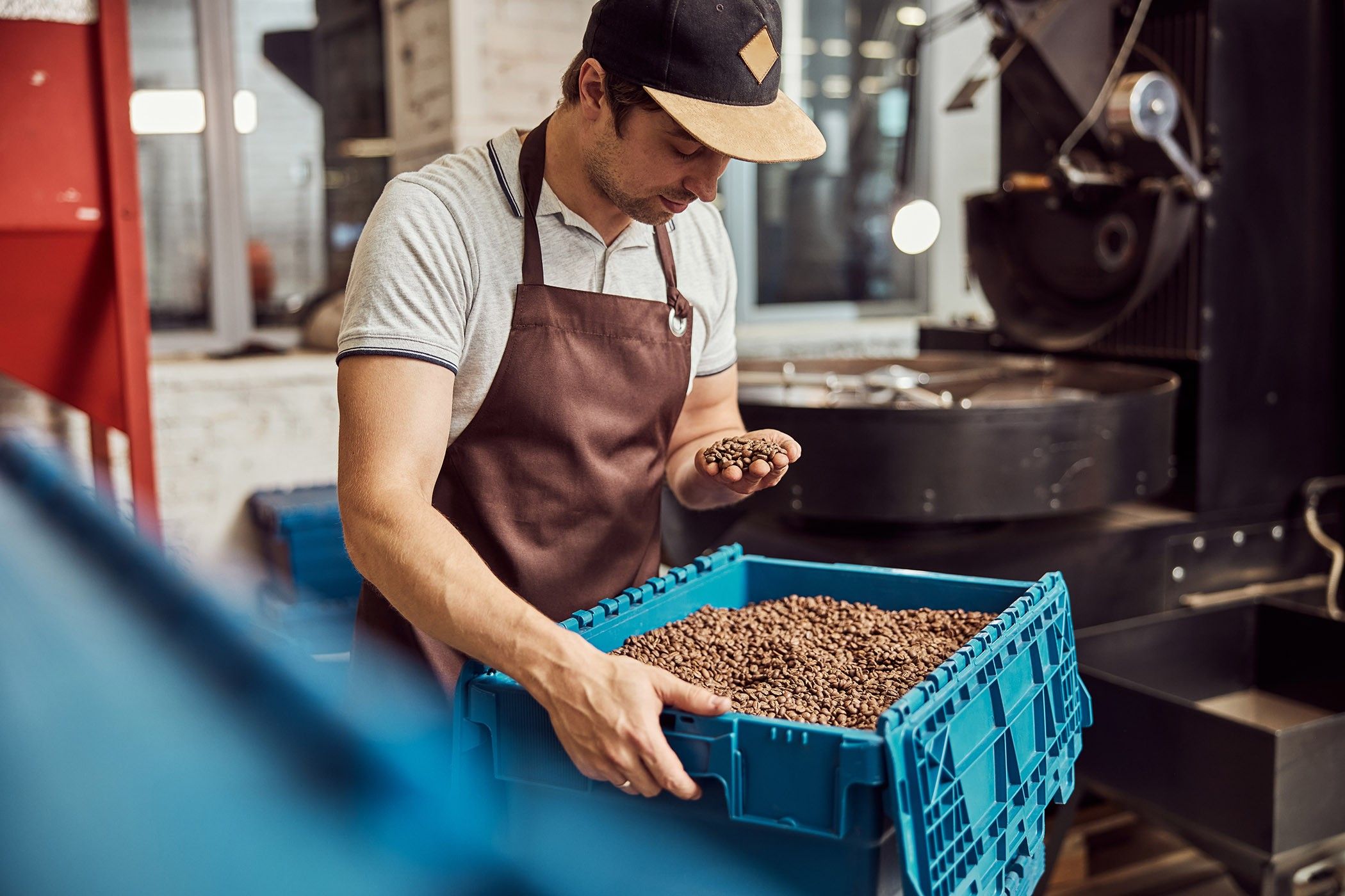 A man holds coffee beans in one hand and a blue bin of coffee beans in the other. 